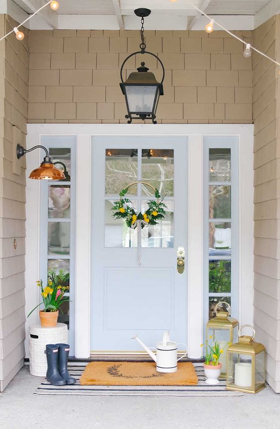 stylish spring porch decor with a watering can, candle lanterns, bright blooms in pots and a bright yellow wreath