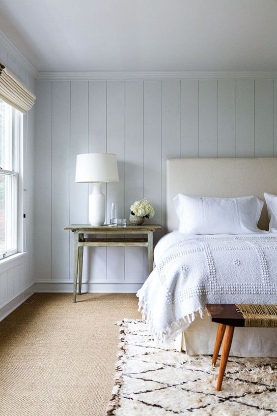 a beautiful coastal bedroom with whitewashed wooden walls, a neutral upholstered bed, vintage benches and tables and neutral bedding