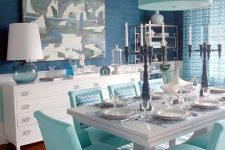 a bold dining room with navy walls and white paneling, white sideboards, a white dining table and turquoise chairs and a pendant lamp