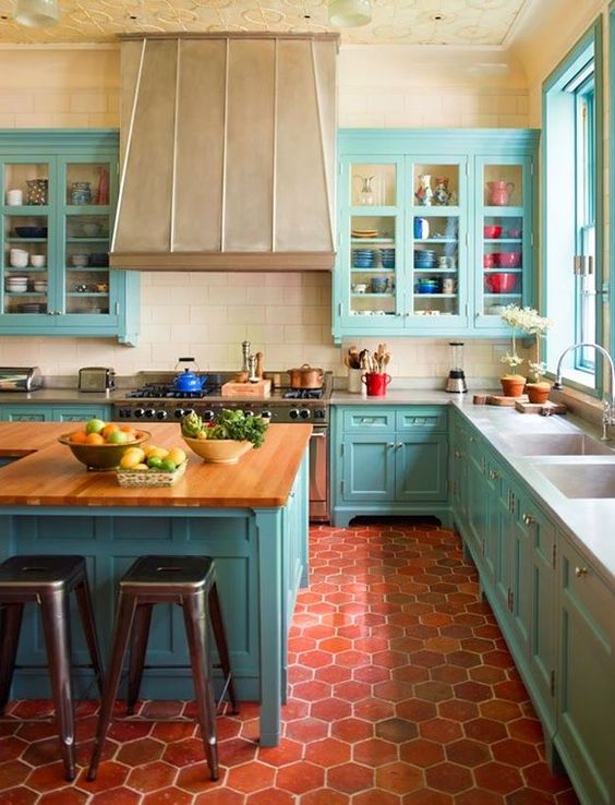 a bright turquoise kitchen with shaker style cabinets, a large metal hood, a large kitchen island with butcherblock countertops and terracotta hexagon tiles on the floor