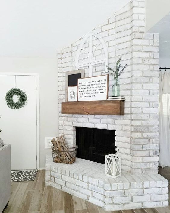 a chic whitewashed brick fireplace with a wooden mantel, artworks and eucalyptus, a wire basket with firewood