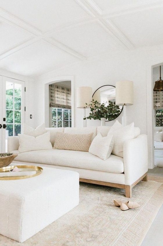 a cool living room with a white sofa and a matching ottoman, with a round mirror, two lamps and some chic accessories