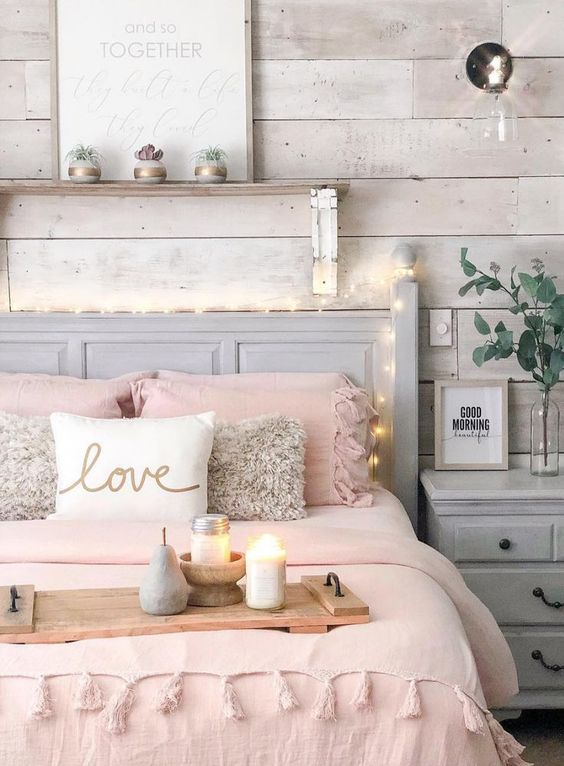 a cozy bedroom with a whitewashed wooden accent wall, neutral furniture, lights, a wall sconce, pink bedding and artworks