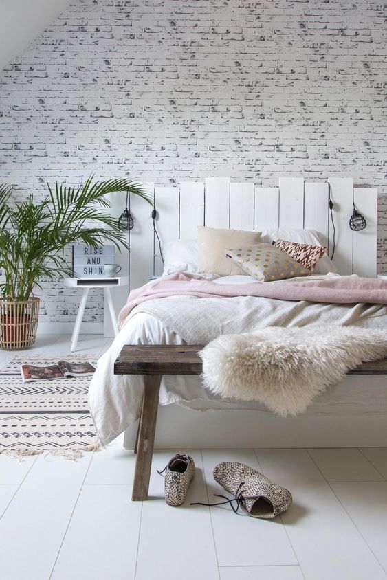 a cozy bedroom with a wooden floor and a whitewashed brick wall, a wooden bed and a bench, catchy bedding and lights