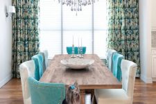 a dining room with a ceiling with a chic crystal chandelier, a wooden dining table, white and turquoise chairs with decorative nails