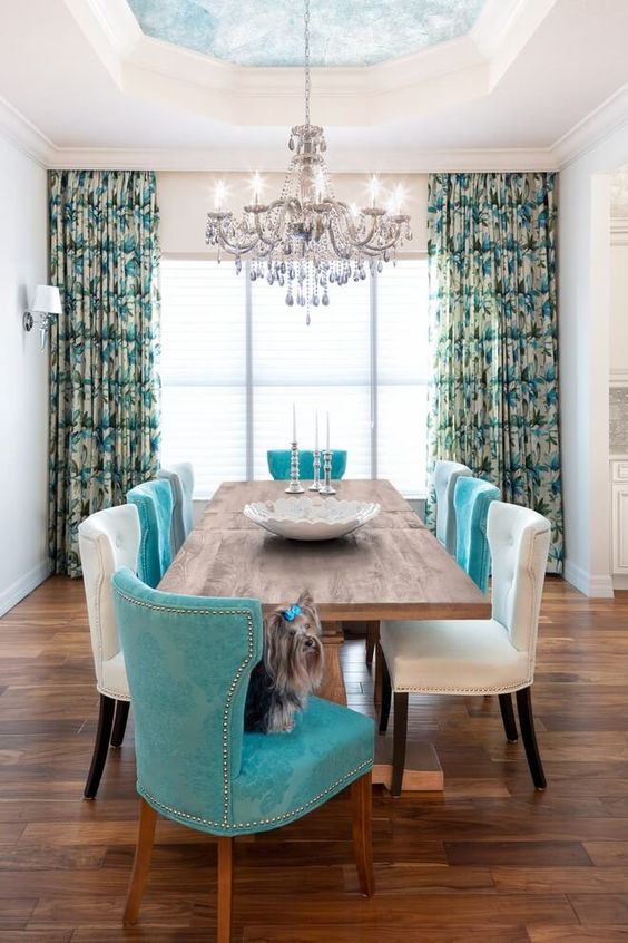 a dining room with a ceiling with a chic crystal chandelier, a wooden dining table, white and turquoise chairs with decorative nails