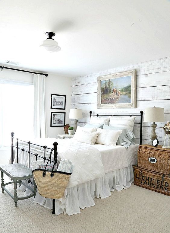 a farmhouse bedorom with a whitewashed wooden wall, a metal bed, stacked woven chests with refined lamps and a rustic artwork