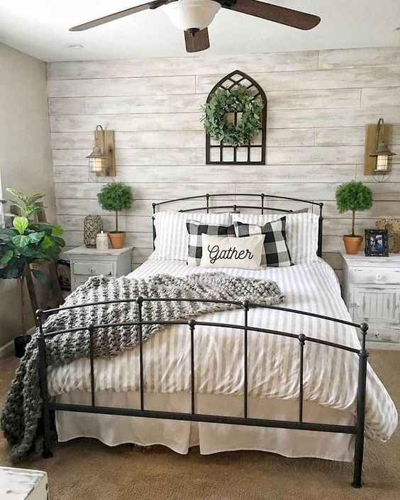 a farmhouse bedroom with a whitewashed wood accent wall, a black metal bed, whitewashed nightstands, potted plants and a greenery wreath