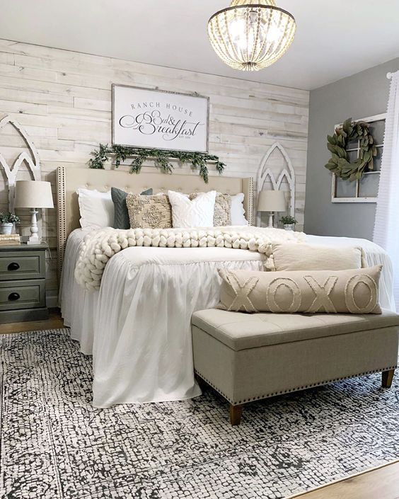 a farmhouse bedroom with a whitewashed wooden wall, neutral upholstered bed and ottoman, vintage nightstands, greenery and a vintage chandelier