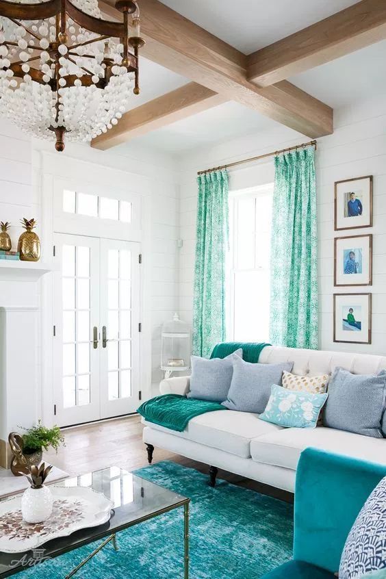 a gorgeous living room with a fireplace, a white sofa, a glass coffee table, wooden beams on the ceiling and turquoise textiles