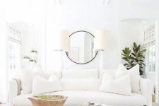 a modern white living room with a soft sofa and an ottoman, table lamps and a potted plant plus potted greenery