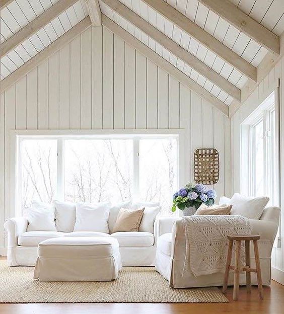 a neutral living room with whitewashed wooden wall and a ceiling, white furniture, some wooden stools and blooms