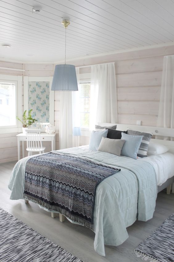 a neutral rustic bedroom with whitewashed wooden walls, simple and chic furniture, blue and grey bedding and a blue pendant lamp, a vanity corner with a mirror