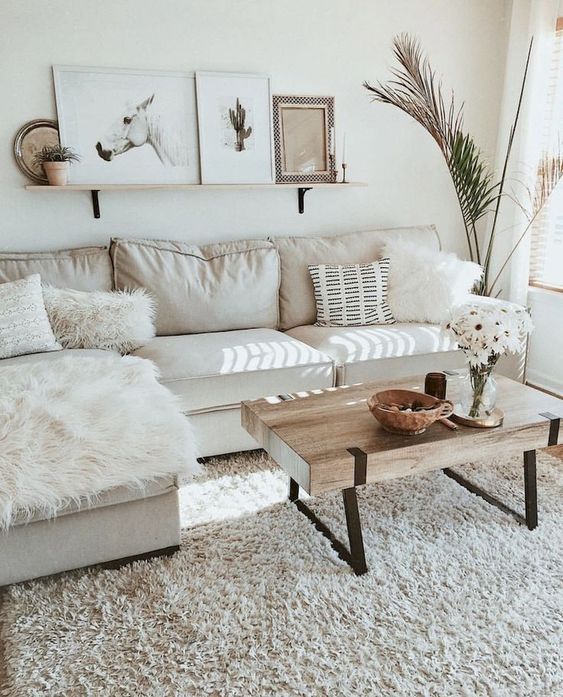 a neutral small living room with a sectional sofa, a wooden table, lots of fur pillows and a gallery wall on a shelf