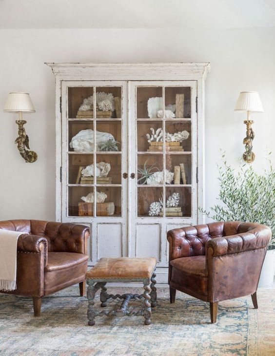 a shabby chic space with a whitewashed display cabinet with glass doors and leather chairs, a leather sotol and wall sconces