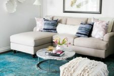 a small colorful living room with a neutral sofa, a turquoise rug, a boho ottoan, pretty plates and an artwork