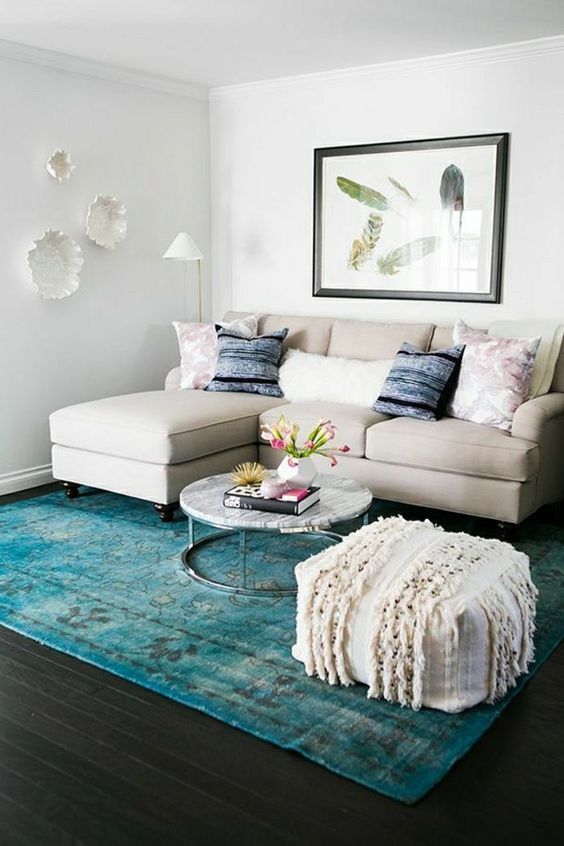 a small colorful living room with a neutral sofa, a turquoise rug, a boho ottoan, pretty plates and an artwork