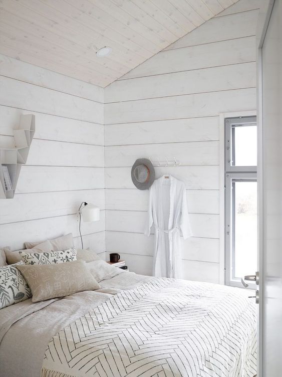 a small neutral bedroom with whitewashed wooden walls, a bed, some sconces and built-in lights is filled with light