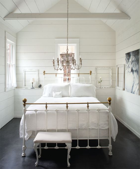 a small yet refined bedroom with whitewashed wooden walls and a ceiling, a metal bed, a sophisticated chandelier and a stool