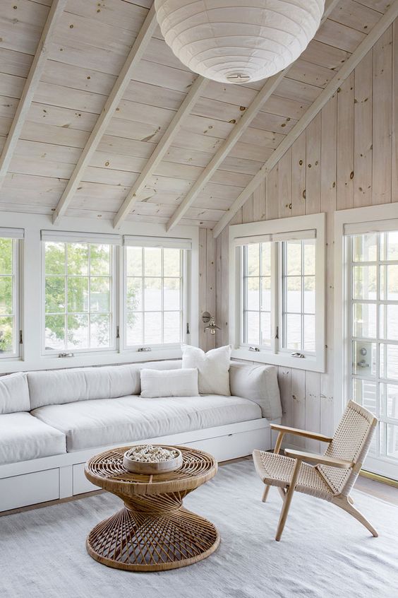 a sunroom with whitewashed wooden walls, a neutral sofa, a woven chair and a rattan coffee table, a pendant paper lamp