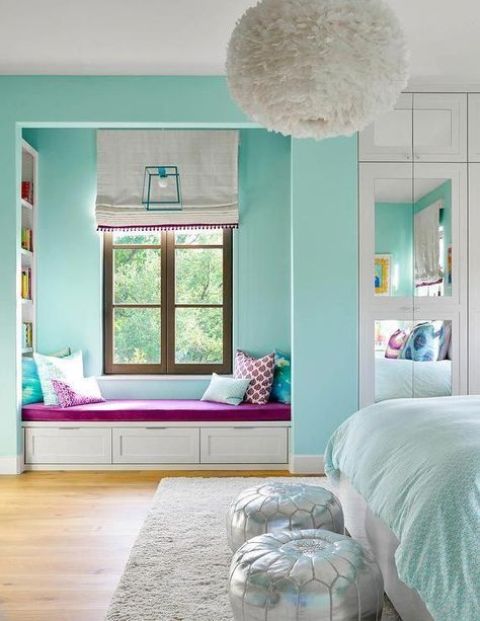 A turquoise bedroom with a window reading nook with built in shelves, a bed with turquoise and white bedding and silver poufs