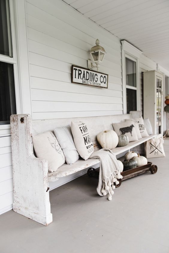 a white farmhouse porch with a whitewashed bench, neutral and printed pillows, a cart with pumpkins, a blanket and a sign