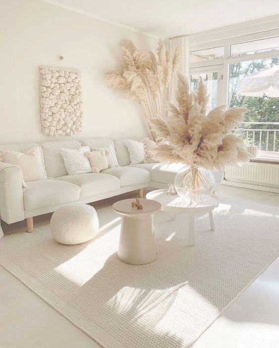 An all white boho living room with a sectional, a pouf, a couple of side tables, pampas grass and a cool wall art