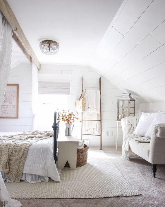 an attic bedroom with whitewashed wooden walls, neutral and cozy furniture, neutral bedding and lace curtains is chic