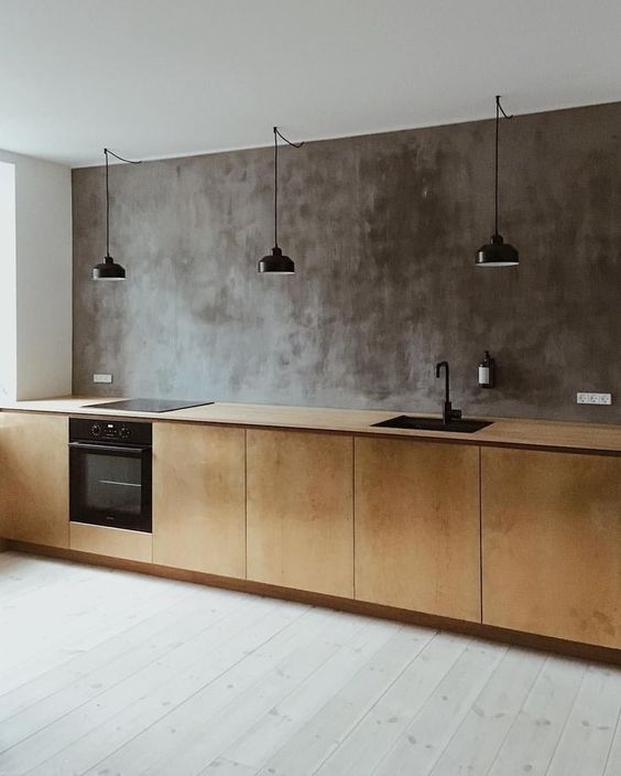 an industrial kitchen with a concrete wall, plywood cabients and a whitewashed floor plus black lamps