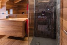 a beautiful chalet bathroom clad with wood, with a shower space done with black faux stone tiles, built-in lights and a floating vanity