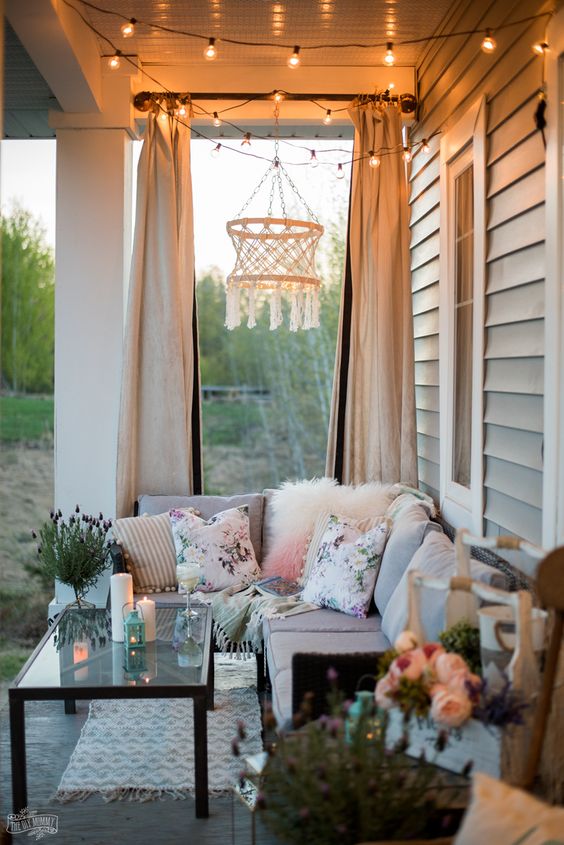 a boho summer porch with a neutral sectional sofa, bright printed pillows, a macrame chandelier, a glass coffee table and candles