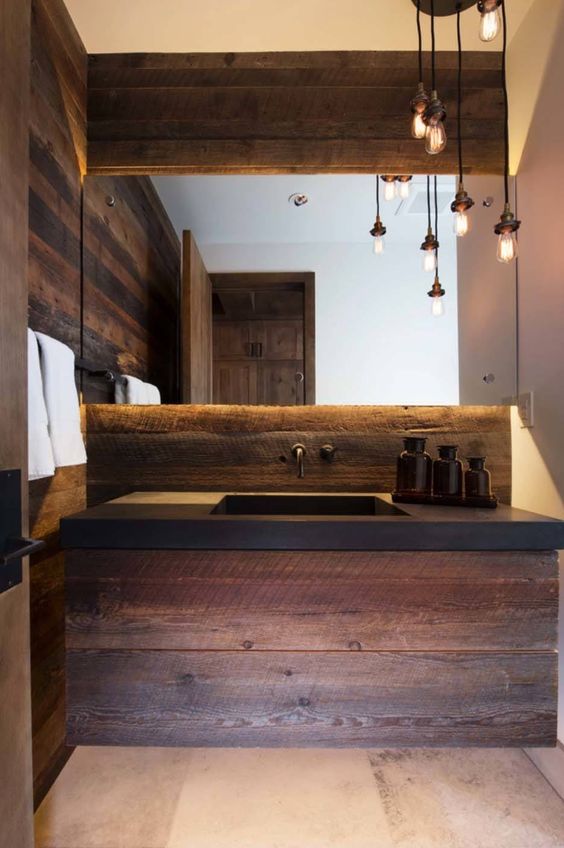 A catchy chalet bathroom done with dark stained textural wood, a black concrete countertop, pendant bulbs and a lit up mirror