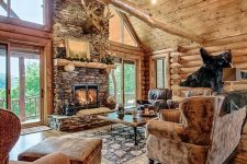a chalet living room with wooden walls, a floor and a ceiling, a stone clad fireplace, leather and fabric upolstered furniture and taxidermy