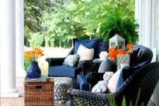 a chic summer porch with navy wicker chairs, a wicker ottoman, potted blooms and greenery and candle lanterns