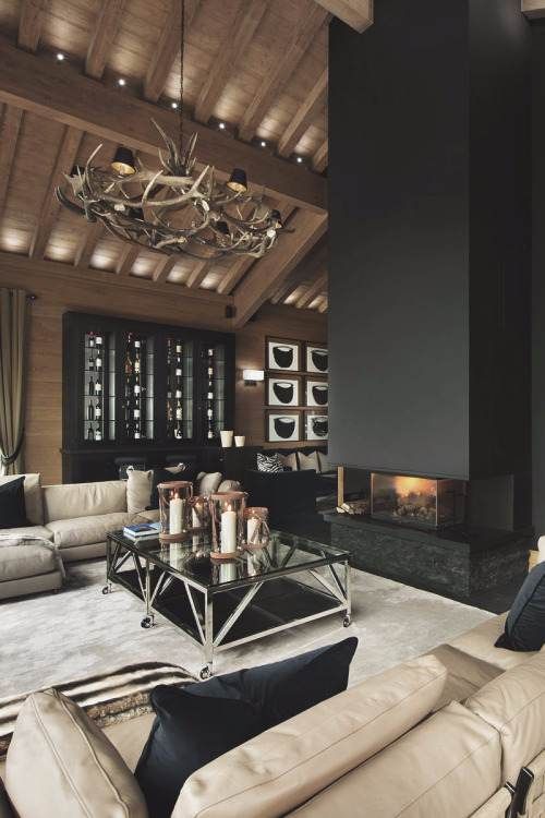 a contemporary chalet living room with modern neutral and vintage black furniture, a glass coffee table, a fireplace and an antler chandelier