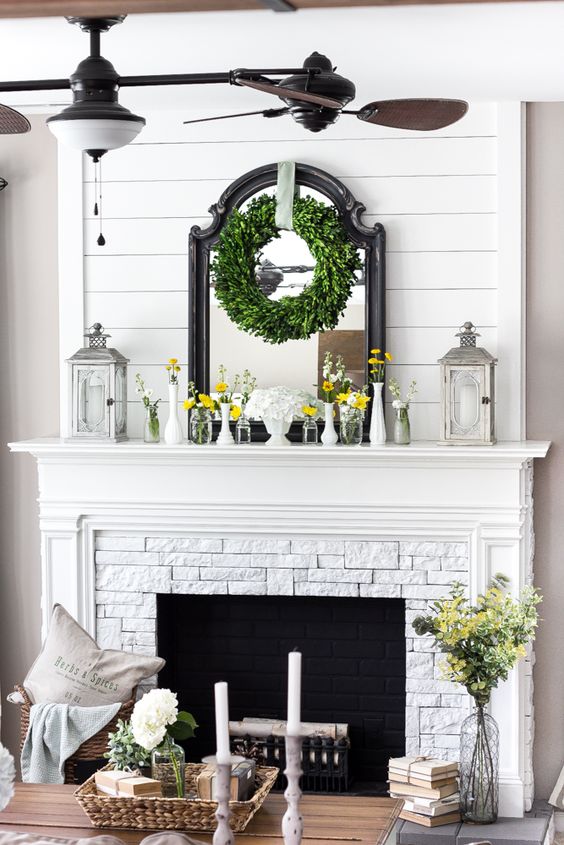 a farmhouse mantel with candle lanterns, vases with bright blooms, a greenery wreath and a mirror