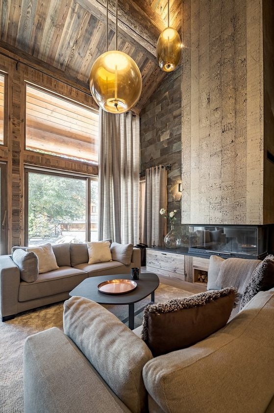 A jaw dropping chalet living room with all wood around, a stone wall, a catchy fireplace, modern furniture and hanging lamps