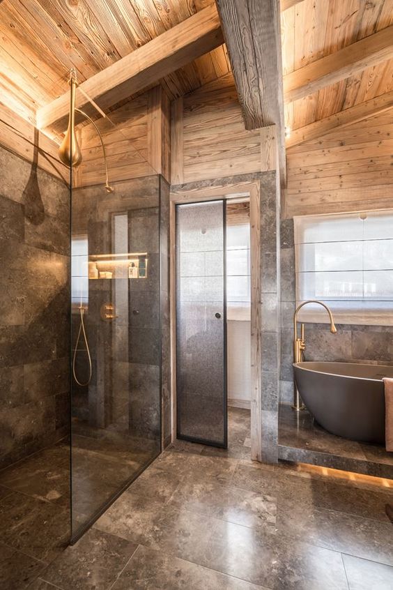 A luxurious chalet bathroom done with grey marble and light stained wood, with brass touches and a bathtub on a platform