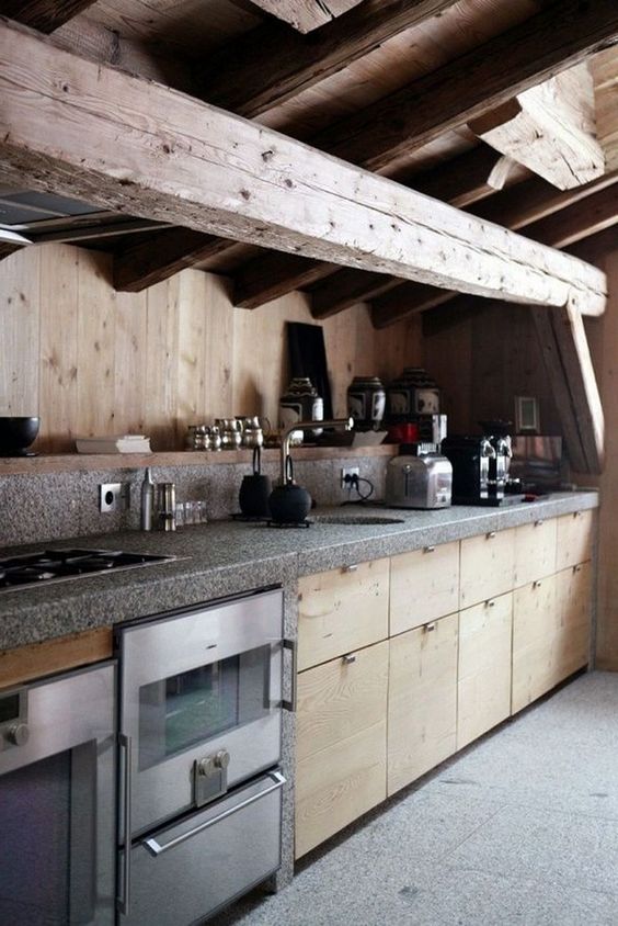 a minimalist chalet kitchen done with wooden beams, sleek wooden cabinets, grey countertops and metal appliances