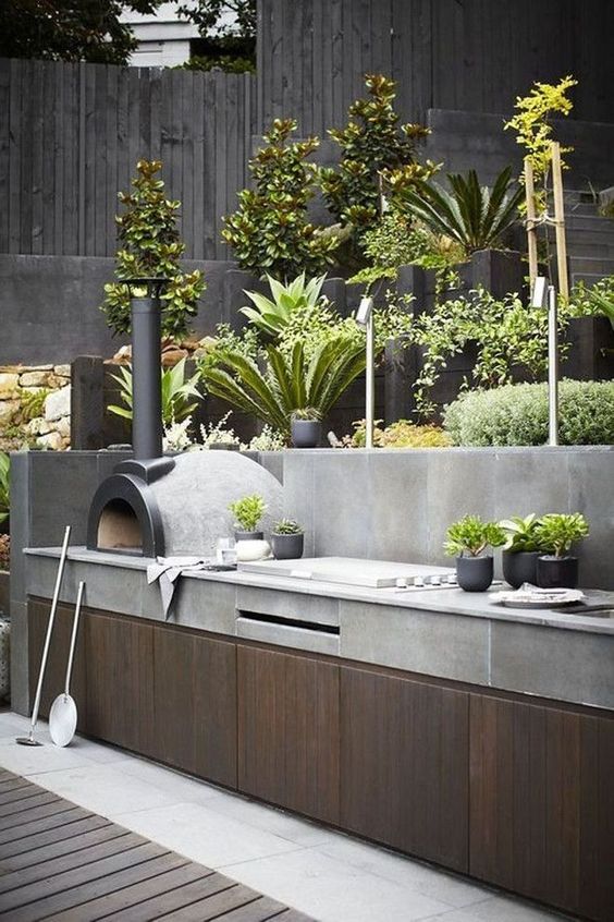 a minimalist outdoor bbq area of wood and concrete, with a grill and a pizza oven plus potted greenery
