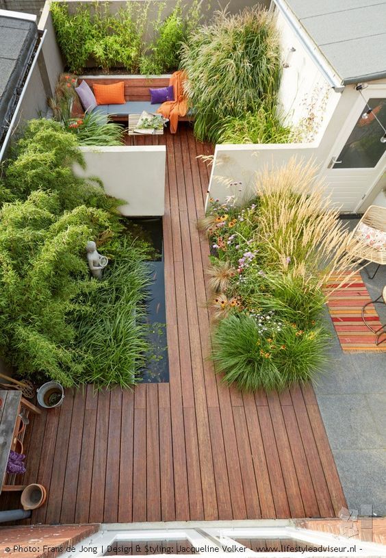 A minimalist townhouse garden with a wooden deck, a pond, planted herbs and grasses and a built in bench