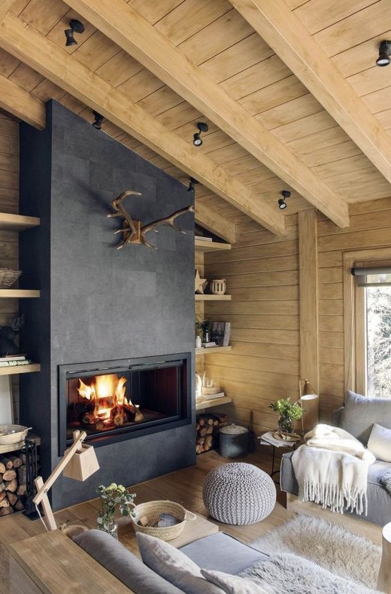 a modern chalet living room with wooden walls and a ceiling, a concrete fireplace, firewood storage, grey sofas