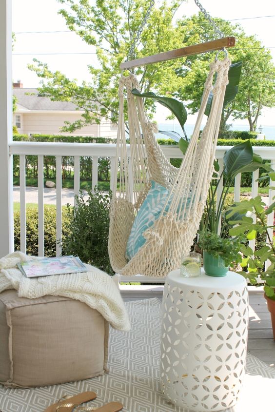 a modern farmhouse porch with a hanging macrame chair, a burlap ottoman, a white side table and greenery