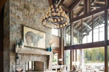 a mountain cabin living room with a wooden ceiling, a stone clad fireplace and stylish neutral furniture plus a statement chandelier
