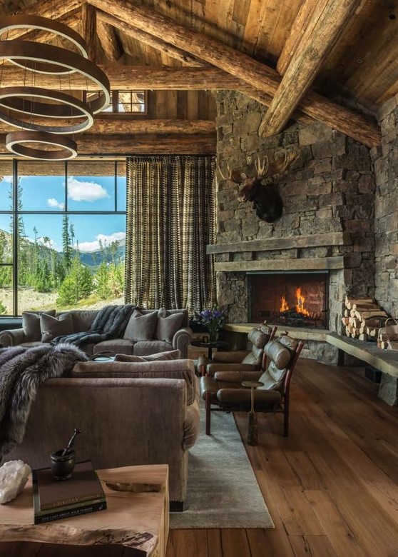 a rustic mountain living room with a wooden ceiling with beams, a stone clad fireplace and cozy furniture plus a catchy chandelier