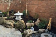 a small Japanese garden with large rocks, a bamboo fountain, pebbles, a stone bowl and trees plus a large stone lantern