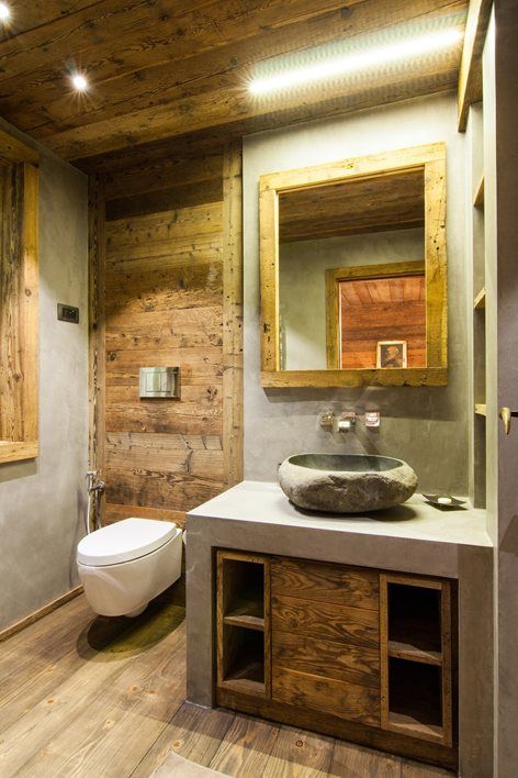 A small chalet bathroom clad with cocnrete and with lots of wood, with a mirror in a wooden frame and a built in vanity