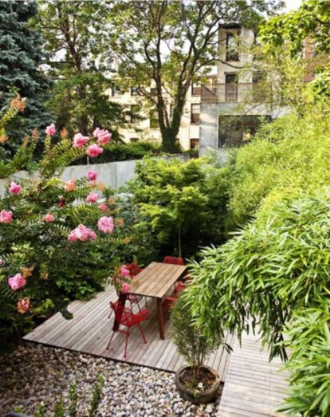 a small townhouse garden with a wooden deck, pebbles, growing greenery and trees plus red chairs and a dining table