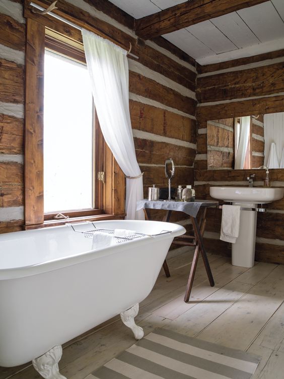 A vintage chalet bathroom with various types of wood, a clawfoot tub, a free standing sink and a trestle table, neutral textiles
