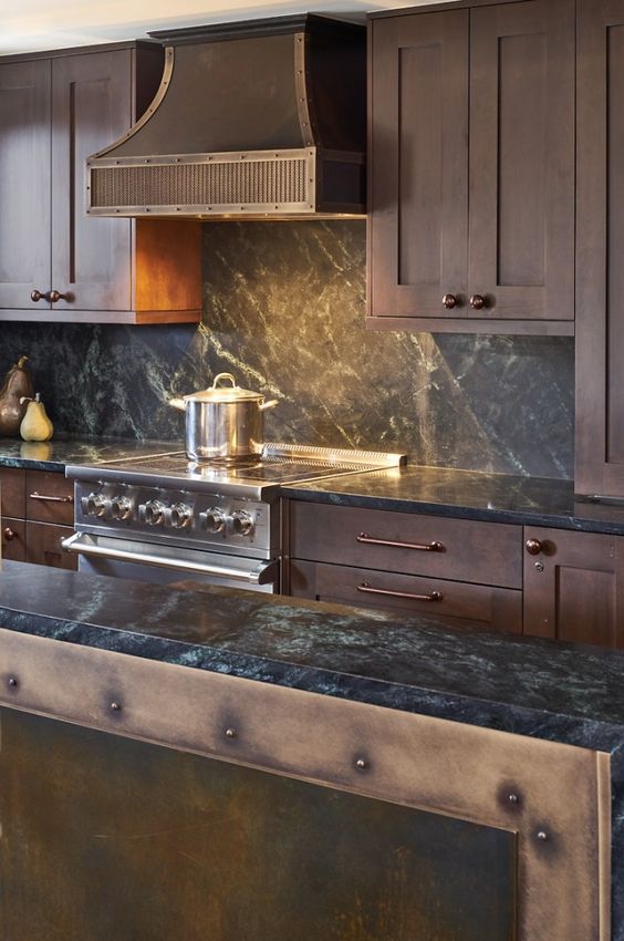 a vintage chalet kitchen with dark stained cabinets, black stone countertops, a vintage metal hood and lights